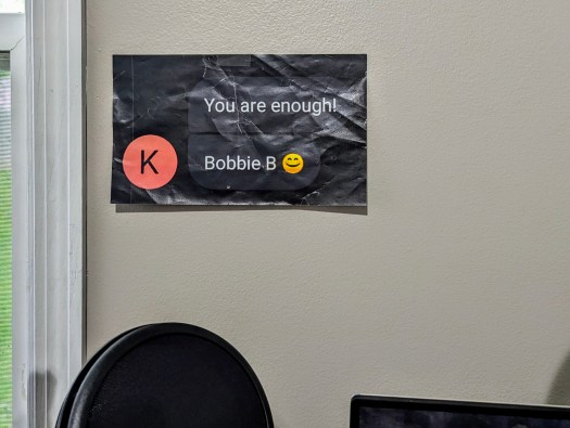 Photo of a wall with a crinkled paper that is a printed screenshot of a text message conversation from sender "K." The first message reads, "You are enough!" The second message reads, "Bobbie B :)"